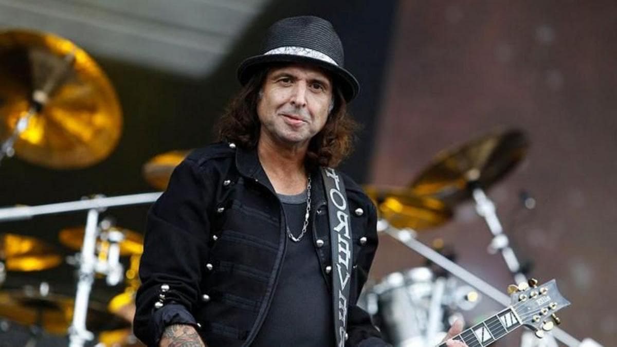 Phil, who has shoulder-length dark brown hair, and is wearing a black bucket hat, black t-shirt and black jacket. He is holding a guitar and is stood on a stage with a large drum set behind him.