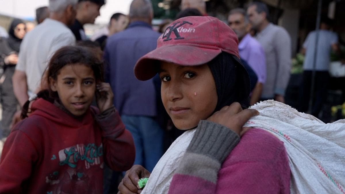 A girl wearing a pink baseball cap with a white sack over her shoulder looks at the camera, with a second girl behind her and a crowd of people behind them standing further away